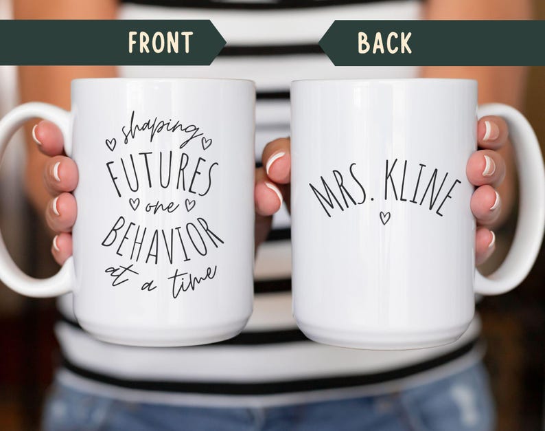 May include: White ceramic mug with black text that reads "shaping FUTURES one BEHAVIOR at a time" on the front and "MRS. KLINE" on the back. The mug has a white handle and is held by a person.