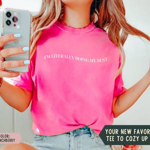 K&ouml;nnte beinhalten: Ein leuchtend pinkfarbenes T-Shirt mit dem wei&szlig;en Text "I'M LITERALLY DOING MY BEST". Das Bild enth&auml;lt auch den Text "YOUR NEW FAVORITE TEE TO COZY UP IN!" und "COLOR: CRUNCHBERRY". Das Shirt hat eine kr&auml;ftige Farbe.