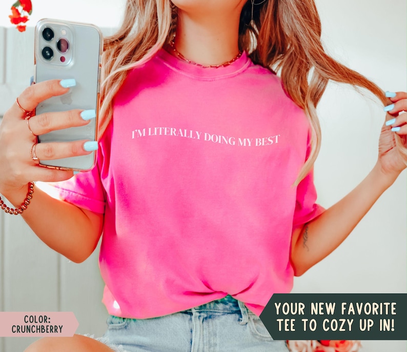 K&ouml;nnte beinhalten: Ein leuchtend pinkfarbenes T-Shirt mit dem wei&szlig;en Text "I'M LITERALLY DOING MY BEST". Das Bild enth&auml;lt auch den Text "YOUR NEW FAVORITE TEE TO COZY UP IN!" und "COLOR: CRUNCHBERRY". Das Shirt hat eine kr&auml;ftige Farbe.