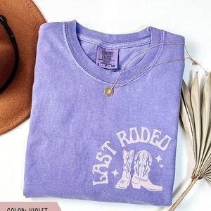 May include: A violet Comfort Colors t-shirt with the text "LAST RODEO" and a graphic of cowboy boots. A gold necklace with a pendant is draped across the shirt. The shirt is folded on a white surface.