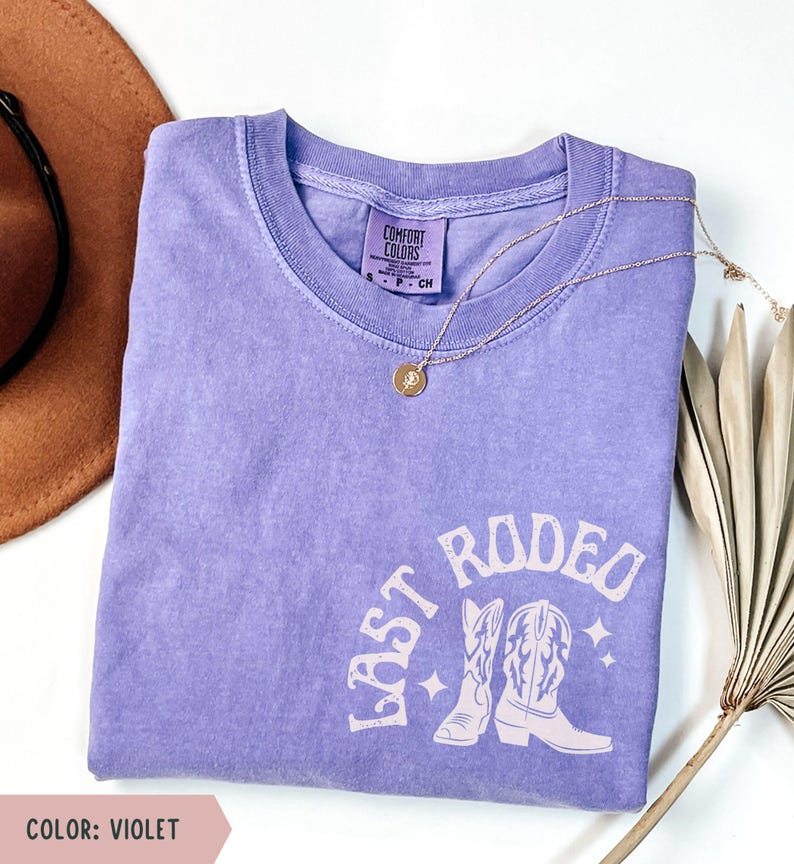 May include: A violet Comfort Colors t-shirt with the text "LAST RODEO" and a graphic of cowboy boots. A gold necklace with a pendant is draped across the shirt. The shirt is folded on a white surface.
