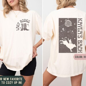 May include: Ivory-colored t-shirt with a rodeo-themed graphic on the front and back. The front features the text "LAST RODEO" with cowboy boots. The back displays "KAYLA'S BACH" with a bull riding design and disco ball.