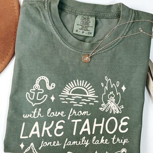 May include: A moss green t-shirt with white graphic text and illustrations. The text reads "with love from LAKE TAHOE jones family lake trip." The design includes an anchor, sun, and campfire. A gold necklace with a pendant is on the shirt.