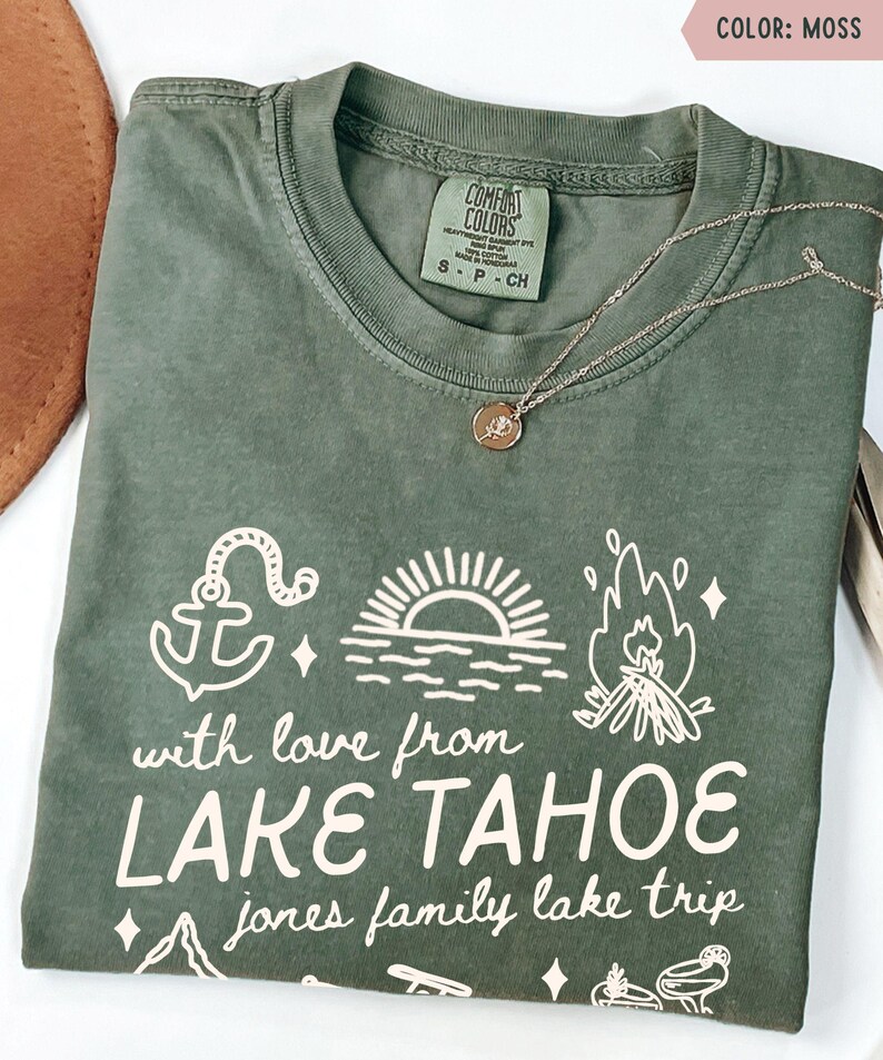 May include: A moss green t-shirt with white graphic text and illustrations. The text reads "with love from LAKE TAHOE jones family lake trip." The design includes an anchor, sun, and campfire. A gold necklace with a pendant is on the shirt.