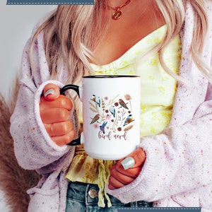 May include: White ceramic mug with a black handle and rim. The mug features a colorful illustration of birds and flowers with the text "bird nerd".