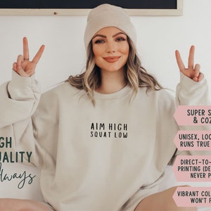 May include: A beige sweatshirt with the text "AIM HIGH SQUAT LOW" printed on the front. The sweatshirt is super soft and cosy, unisex, and runs true to size. It is made with direct-to-garment printing, so the design will never peel. The sweatshirt is available in vibrant colours that won't fade.
