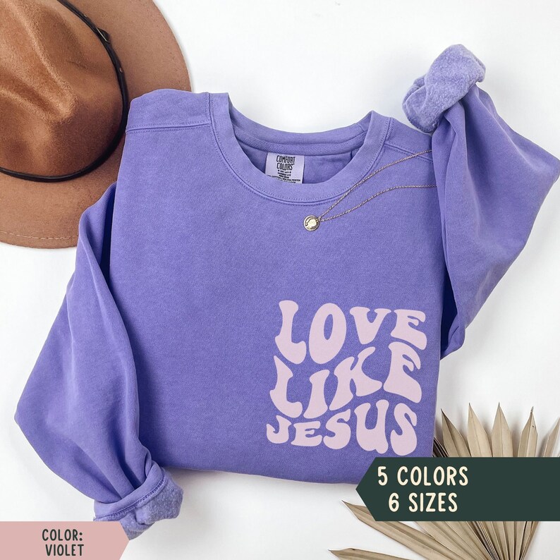 K&ouml;nnte beinhalten: Lilafarbener Pullover mit dem Text "LOVE LIKE JESUS" in rosafarbenen Retro-Lettern. Der Pullover ist in 5 Farben und 6 Gr&ouml;&szlig;en erh&auml;ltlich.