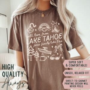 May include: Espresso-colored t-shirt with a white graphic design featuring an anchor, lake scene, campfire, trees, boat, coffee cup, and sunglasses. The text reads "with love from LAKE TAHOE jones family lake trip". The words "HIGH QUALITY Always" are also visible.