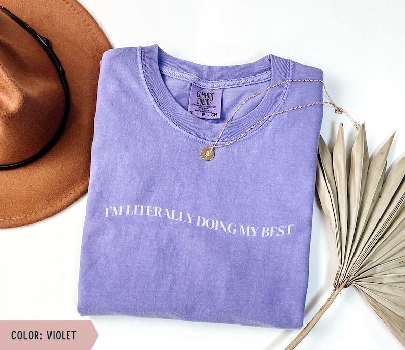 K&ouml;nnte beinhalten: Ein violettes Comfort Colors T-Shirt mit dem wei&szlig;en Aufdruck "I'M LITERALLY DOING MY BEST". Eine goldene Halskette und ein brauner Hut sind ebenfalls im Bild. Das Shirt ist Gr&ouml;&szlig;e S.