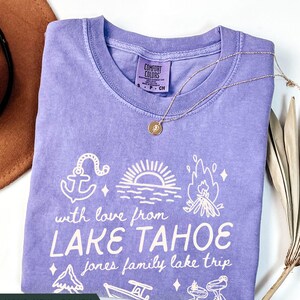 May include: A violet Comfort Colors t-shirt with a white graphic design featuring an anchor, sunset, campfire, and other lake-themed illustrations. The text reads "with love from LAKE TAHOE" and "jones family lake trip". A gold necklace is draped across the shirt.