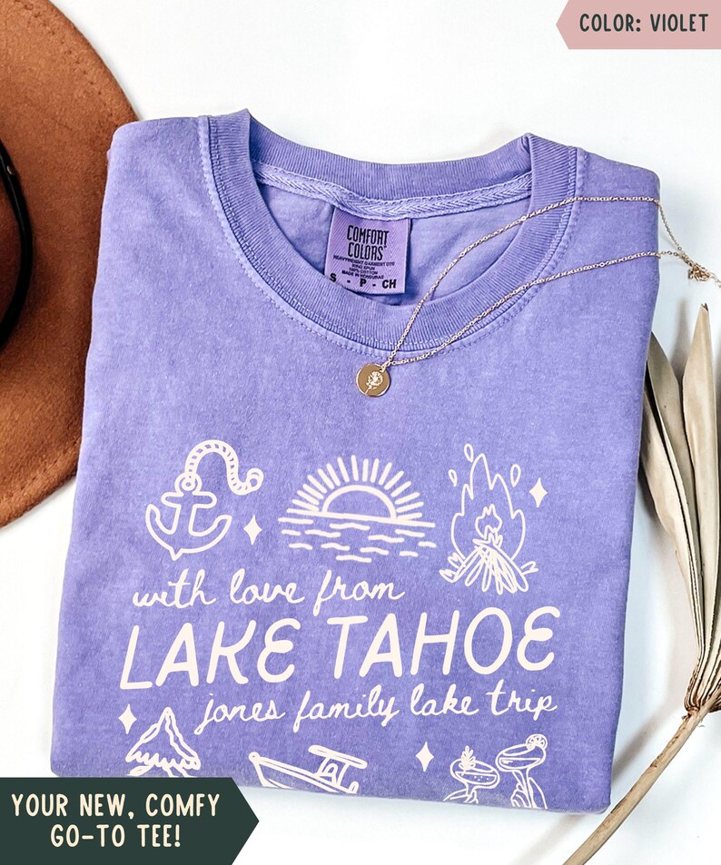 May include: A violet Comfort Colors t-shirt with a white graphic design featuring an anchor, sunset, campfire, and other lake-themed illustrations. The text reads "with love from LAKE TAHOE" and "jones family lake trip". A gold necklace is draped across the shirt.