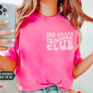 May include: A bright pink t-shirt with the text "2ND GRADE TEACHER CLUB" in a retro font. The shirt also has the text "YOUR NEW FAVORITE TEE TO COZY UP IN!" and the color "CRUNCHBERRY".