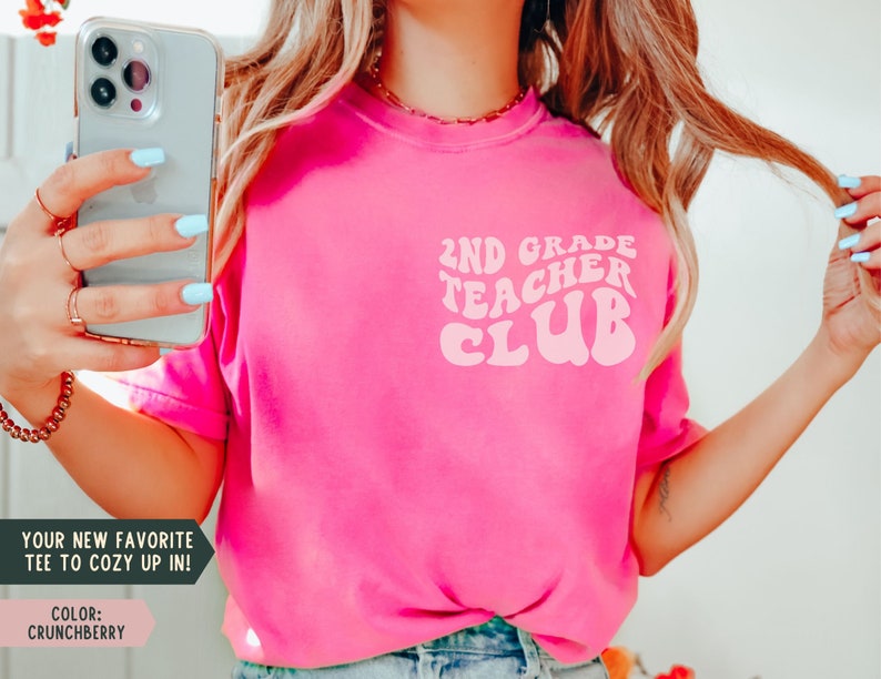 May include: A bright pink t-shirt with the text "2ND GRADE TEACHER CLUB" in a retro font. The shirt also has the text "YOUR NEW FAVORITE TEE TO COZY UP IN!" and the color "CRUNCHBERRY".
