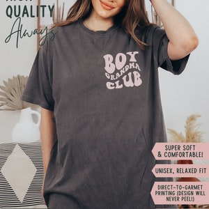 May include: A dark grey t-shirt with a pink graphic that says "Boy Grandma Club". The shirt is unisex and has a relaxed fit. The text "Color: Pepper" is visible at the bottom of the image.
