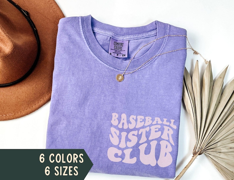 May include: A lavender-colored t-shirt with the text "BASEBALL SISTER CLUB" in a retro font. The shirt has a crew neck and short sleeves. A gold necklace with a pendant is draped on the shirt. The image also shows a brown hat and a sprig of dried leaves.