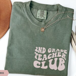 May include: A moss green t-shirt with the text "2ND GRADE TEACHER CLUB" in a retro font. A gold necklace with a pendant and a brown hat are also in the image. The shirt is made of a soft material.