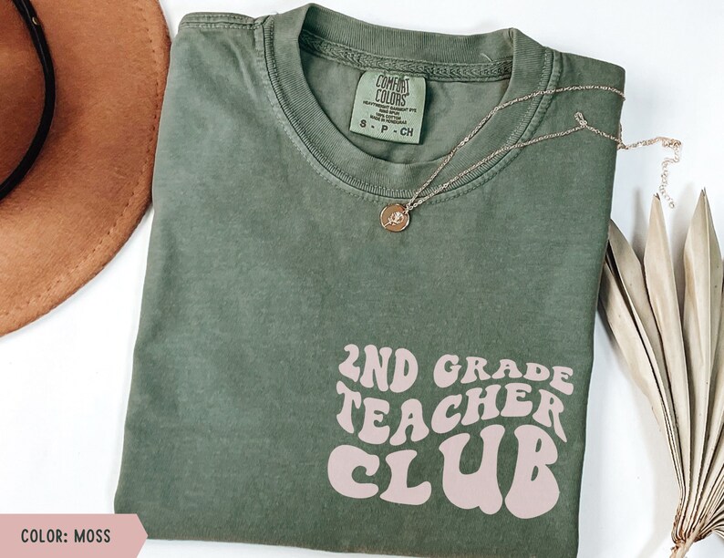 May include: A moss green t-shirt with the text "2ND GRADE TEACHER CLUB" in a retro font. A gold necklace with a pendant and a brown hat are also in the image. The shirt is made of a soft material.