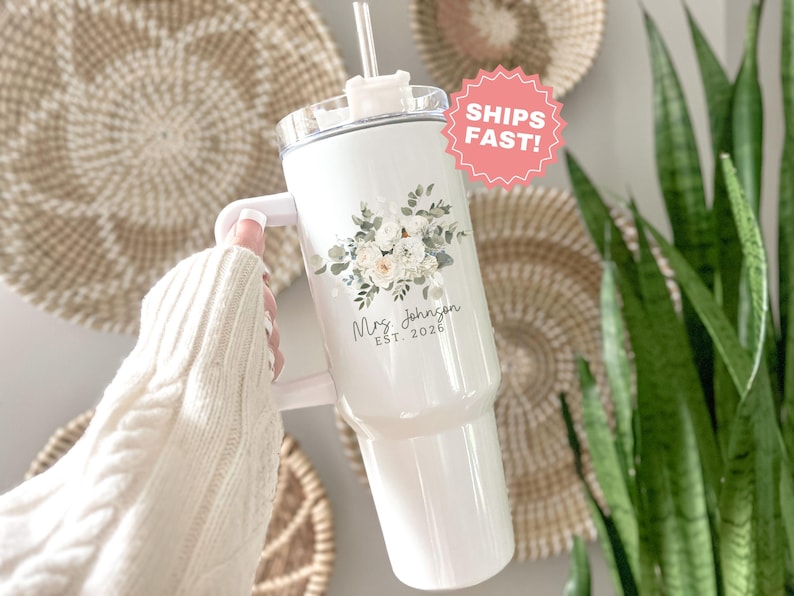 Puede incluir: Vaso blanco con asa, dise&ntilde;o floral y el texto "Mrs. Johnson EST. 2026". El vaso tiene una tapa transparente y una pajita. El dise&ntilde;o incluye flores blancas y verde claro. El vaso lo sostiene una persona que lleva un su&eacute;ter blanco.