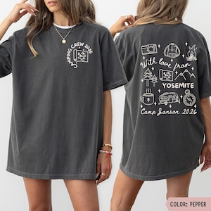 K&ouml;nnte beinhalten: Dunkelgraues T-Shirt mit wei&szlig;en Camping-Motiven. Auf der Vorderseite steht "Camping Crew 2026" und eine Karte. Auf der R&uuml;ckseite befinden sich Illustrationen und der Text: "With love from Yosemite, Camp Janson 2026."