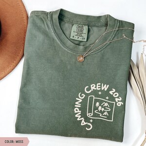 K&ouml;nnte beinhalten: Ein moosgr&uuml;nes T-Shirt mit dem wei&szlig;en Aufdruck "CAMPING CREW 2026". Das Design beinhaltet eine Karte mit B&auml;umen und Bergen. Eine goldene Halskette mit Anh&auml;nger befindet sich auf dem T-Shirt. Das T-Shirt ist gefaltet.