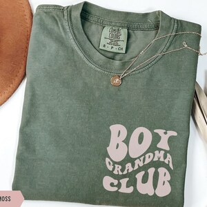 May include: A moss green t-shirt with a pink graphic that says "Boy Grandma Club" in a retro font.