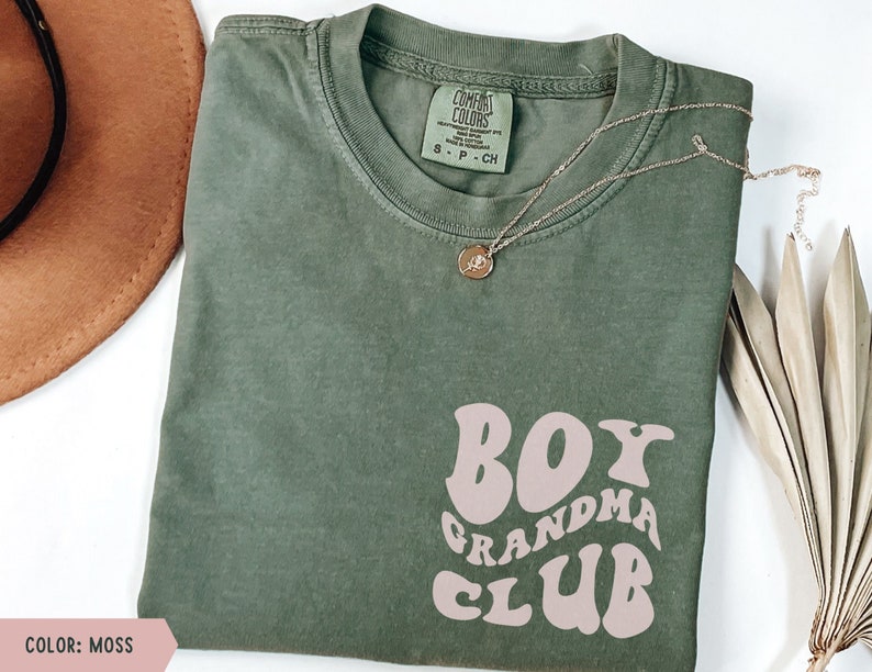 May include: A moss green t-shirt with a pink graphic that says "Boy Grandma Club" in a retro font.