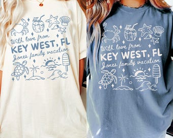 May include: Two oversized t-shirts, one cream and one blue, featuring a Key West vacation design. The design includes illustrations of a crab, coconut drink, starfish, seashell, turtle, palm tree, sun, waves, and an ice pop. The text reads "With love from KEY WEST, FL Jones family vacation."