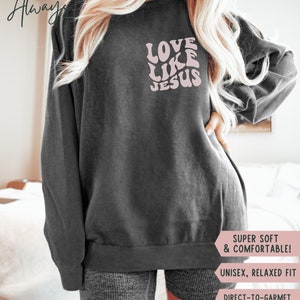 K&ouml;nnte beinhalten: Ein graues Sweatshirt mit dem Text "LOVE LIKE JESUS" in Pink auf der Vorderseite. Das Sweatshirt ist ein Unisex-Modell mit lockerer Passform und aus superweichem und bequemem Stoff gefertigt. Der Text ist in einer Retro-Schriftart mit Wellen.