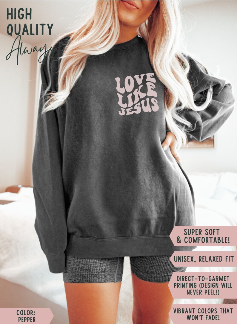 K&ouml;nnte beinhalten: Ein graues Sweatshirt mit dem Text "LOVE LIKE JESUS" in Pink auf der Vorderseite. Das Sweatshirt ist ein Unisex-Modell mit lockerer Passform und aus superweichem und bequemem Stoff gefertigt. Der Text ist in einer Retro-Schriftart mit Wellen.
