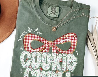 May include: Sage green t-shirt with the words "head baker" and "Cookie Champ!" in a festive design. The design includes a red and white gingham bow with gold accents. A silver necklace with a pendant is also visible.