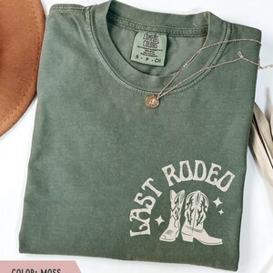 May include: An olive green t-shirt with the words "LAST RODEO" and a graphic of cowboy boots. The shirt has a crew neck and short sleeves. A gold necklace with a pendant is draped across the shirt.