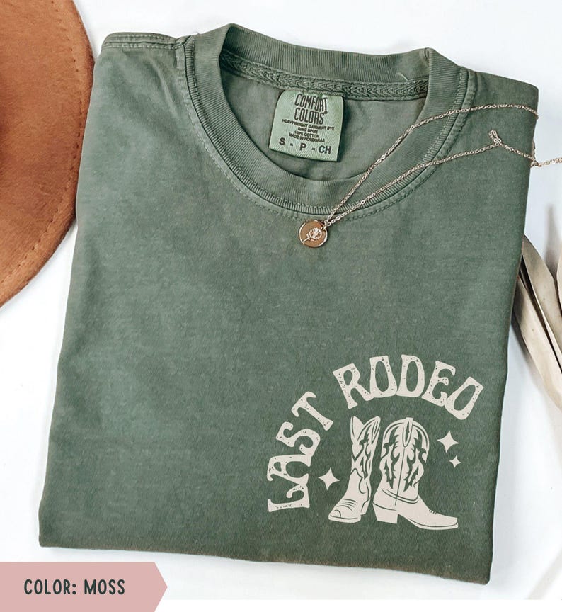 May include: An olive green t-shirt with the words "LAST RODEO" and a graphic of cowboy boots. The shirt has a crew neck and short sleeves. A gold necklace with a pendant is draped across the shirt.
