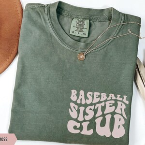 May include: A moss green t-shirt with the text "BASEBALL SISTER CLUB" in a retro font. A gold necklace with a pendant rests on the shirt. The shirt is folded and displayed on a white surface.