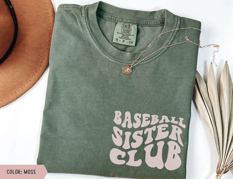 May include: A moss green t-shirt with the text "BASEBALL SISTER CLUB" in a retro font. A gold necklace with a pendant rests on the shirt. The shirt is folded and displayed on a white surface.