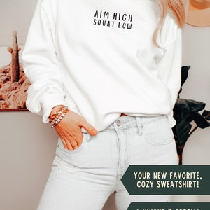 May include: White crew neck sweatshirt with the text "AIM HIGH SQUAT LOW" in black lettering. The sweatshirt is being worn by a person with light blue jeans.
