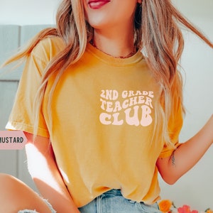 May include: Mustard-colored t-shirt with the text "2ND GRADE TEACHER CLUB" in a retro font. The shirt is a solid color and appears to be made of a soft material. The text is in a light peach color.