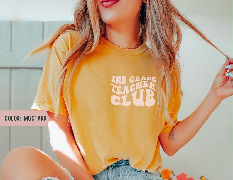 May include: Mustard-colored t-shirt with the text "2ND GRADE TEACHER CLUB" in a retro font. The shirt is a solid color and appears to be made of a soft material. The text is in a light peach color.