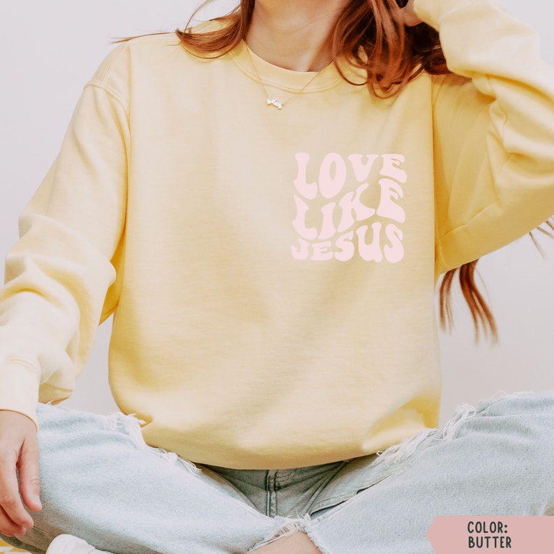 K&ouml;nnte beinhalten: Ein gelber Pullover mit dem Text "LOVE LIKE JESUS" in rosafarbenen Retro-Lettern.