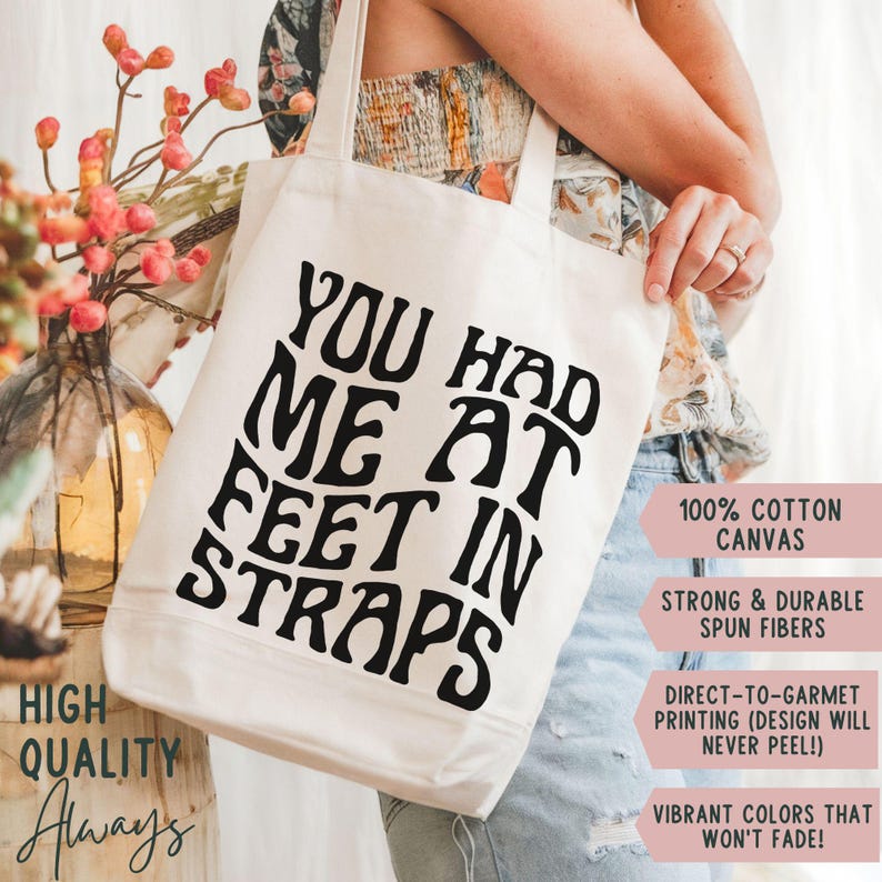 Puede incluir: Bolso tote de lona color crema con el texto "YOU HAD ME AT FEET IN STRAPS" en letras negras en negrita. La bolsa tambi&eacute;n presenta el texto "HIGH QUALITY Always" y detalles sobre el material y la impresi&oacute;n.
