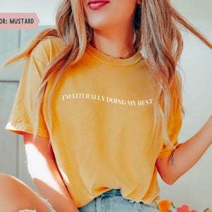K&ouml;nnte beinhalten: Senffarbenes T-Shirt mit dem wei&szlig;en Text "I'M LITERALLY DOING MY BEST". Das Shirt hat kurze &Auml;rmel und einen Rundhalsausschnitt. Das Model tr&auml;gt eine blaue Jeans.
