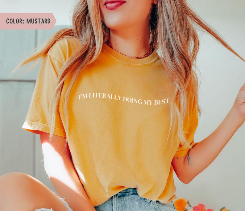 K&ouml;nnte beinhalten: Senffarbenes T-Shirt mit dem wei&szlig;en Text "I'M LITERALLY DOING MY BEST". Das Shirt hat kurze &Auml;rmel und einen Rundhalsausschnitt. Das Model tr&auml;gt eine blaue Jeans.