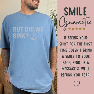 May include: A light blue t-shirt with the text "BUT DID WE SINK?" and an anchor graphic. The text is in a white distressed font. The t-shirt is being worn by a person. The image also includes a pink background with the text "SMILE Guarantee" and five stars.