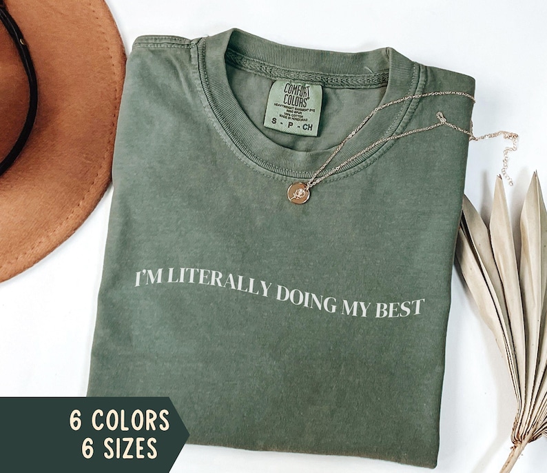 K&ouml;nnte beinhalten: Ein olivgr&uuml;nes T-Shirt mit dem wei&szlig;en Aufdruck "I'M LITERALLY DOING MY BEST". Eine goldene Halskette mit Anh&auml;nger liegt auf dem Shirt. Das Bild zeigt auch einen braunen Hut und Text, der sechs Farben und sechs Gr&ouml;&szlig;en angibt.
