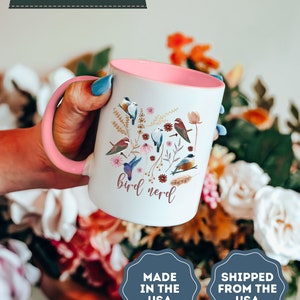 May include: White ceramic mug with a pink rim and handle. The mug features a colorful illustration of birds and flowers with the text "bird nerd".