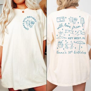 Puede incluir: Camiseta color crema con gráficos azules. La parte delantera presenta el texto "Emma's 30th Birthday" con diseños de estrellas de mar y conchas. La parte trasera tiene un tema de Key West con una cámara, gafas de sol y otras ilustraciones.