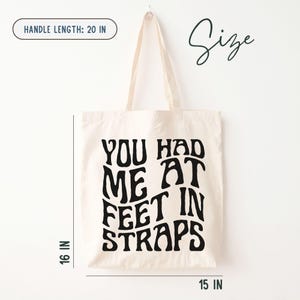 Puede incluir: Bolso tote color crema con texto negro "YOU HAD ME AT FEET IN STRAPS". El bolso mide 40,6 cm de alto, 38,1 cm de ancho y tiene una longitud de asa de 50,8 cm.