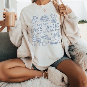 May include: Ivory-colored sweatshirt with a blue graphic that reads "With love from Lake Tahoe" and includes illustrations of an anchor, trees, a boat, and sunglasses. The shirt is a crew neck and has long sleeves.