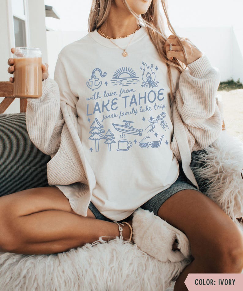 May include: Ivory-colored sweatshirt with a blue graphic that reads "With love from Lake Tahoe" and includes illustrations of an anchor, trees, a boat, and sunglasses. The shirt is a crew neck and has long sleeves.