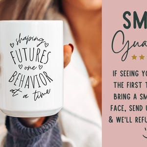 May include: White ceramic mug with the text "shaping FUTURES one BEHAVIOR at a time" in black script. The mug is held by a person. A pink background with the words "SMILE Guarantee" and a refund policy is visible.