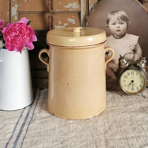 Pode incluir: Uma natureza morta vintage com um pote amarelo com tampa, um jarro branco com flores cor-de-rosa e um retrato em sépia. Persianas de madeira desgastadas e um pequeno despertador completam a cena. O pote tem cerca de 25 cm de altura.